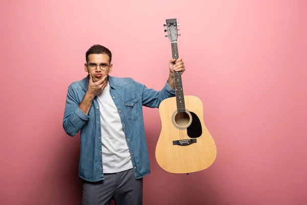 Bell'uomo che sorride alla macchina fotografica e tiene la chitarra acustica su sfondo rosa — Foto stock