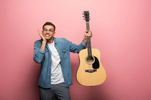 Joven hombre sonriente haciendo muecas mientras sostiene la guitarra acústica sobre fondo rosa - foto de stock