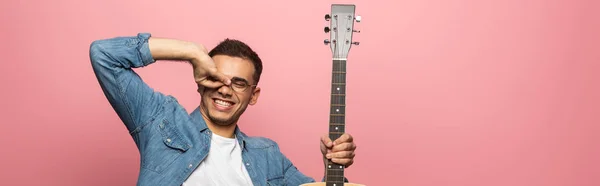 Panoramische Aufnahme eines lächelnden Mannes mit Akustikgitarre, der isoliert auf pink gestikuliert — Stockfoto