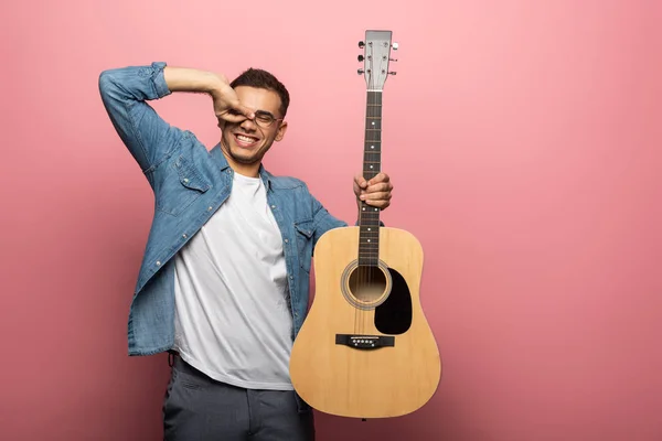 Homem sorridente com guitarra acústica gesticulando sobre fundo rosa — Fotografia de Stock