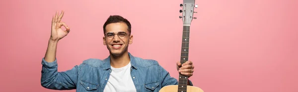 Panoramic shot of man with acoustic guitar smiling at camera and showing okay isolated on pink — Stock Photo