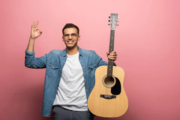 Joven hombre sonriendo a la cámara mientras muestra signo de bien y la celebración de la guitarra acústica sobre fondo rosa - foto de stock