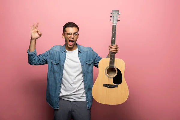 Homem com guitarra acústica piscando e mostrando sinal ok no fundo rosa — Fotografia de Stock