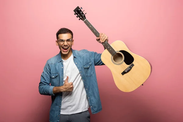 Homme joyeux avec guitare acoustique montrant pouce vers le haut signe sur fond rose — Photo de stock