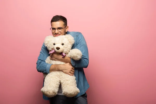 Schöner lächelnder Mann umarmt Teddybär und schaut in die Kamera auf rosa Hintergrund — Stockfoto