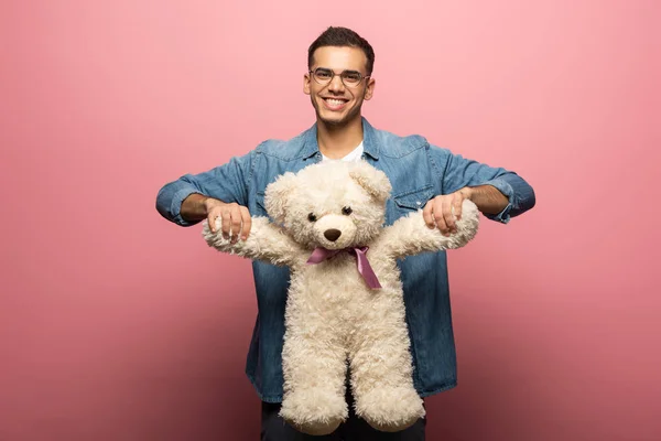 Schöner Mann hält Teddybär in der Hand und lächelt in die Kamera auf rosa Hintergrund — Stockfoto