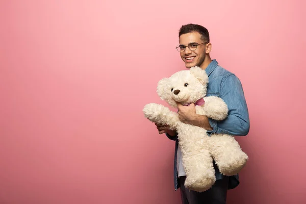 Young man smiling at camera and holding teddy bear on pink background — Stock Photo