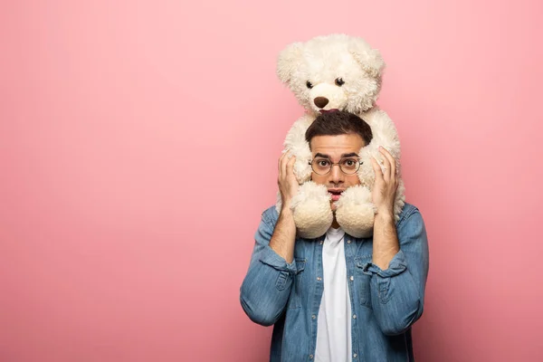 Joven asustado sosteniendo oso de peluche y mirando a la cámara sobre fondo rosa - foto de stock