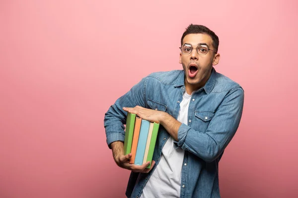 Шокированный человек смотрит в камеру, держа в руках красочные книги на розовом фоне — стоковое фото
