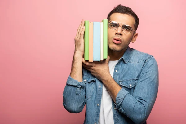 Thoughtful young man looking at camera while holding books on pink background — Stock Photo