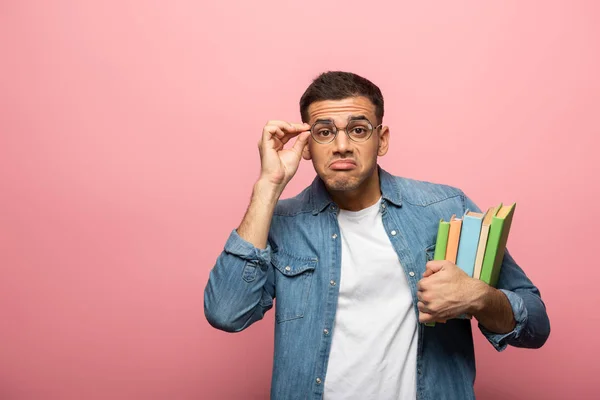 Young man grimacing while adjusting eyeglasses and holding books on pink background — Stock Photo