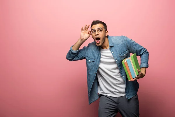 Hombre sorprendido ajustando anteojos y sosteniendo libros de colores sobre fondo rosa - foto de stock