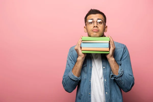 Verwirrter junger Mann hält bunte Bücher in der Hand und blickt in die Kamera auf rosa Hintergrund — Stockfoto
