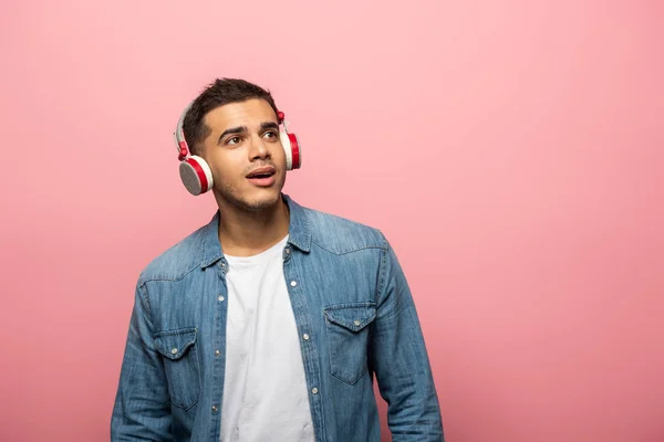 Jovem sonhador em fones de ouvido sem fio olhando para longe isolado em rosa — Fotografia de Stock