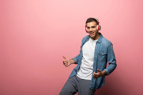 Jeune homme souriant dans les écouteurs montrant pouce vers le haut signe sur fond rose — Photo de stock