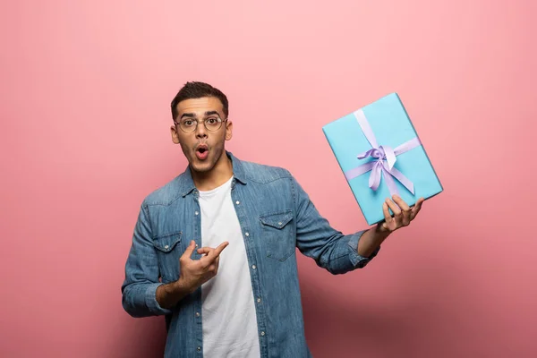 Überraschter Mann blickt in Kamera und zeigt auf Geschenk auf rosa Hintergrund — Stockfoto