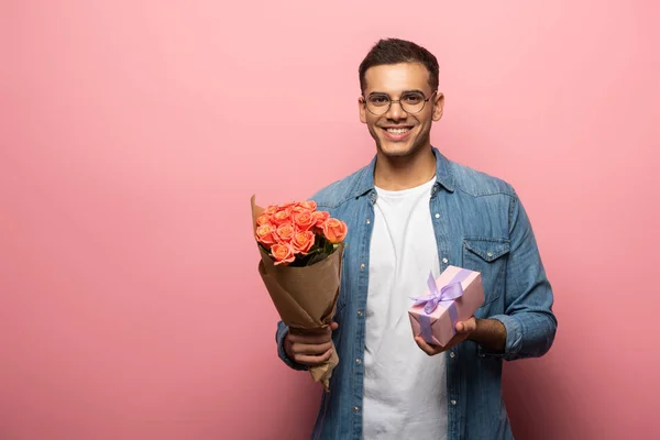 Jovem com buquê e presente sorrindo para a câmera no fundo rosa — Fotografia de Stock