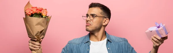 Panoramic shot of pensive man looking at bouquet and holding gift isolated on pink — Stock Photo