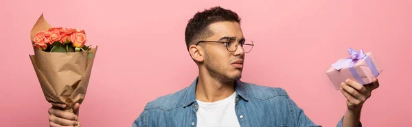 Foto panorámica de un joven mirando el regalo mientras sostiene el ramo aislado en rosa - foto de stock