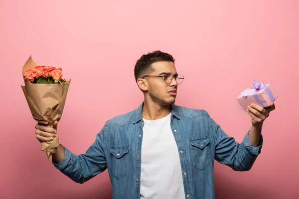 Homme pensif regardant boîte cadeau tout en tenant des fleurs sur fond rose — Photo de stock