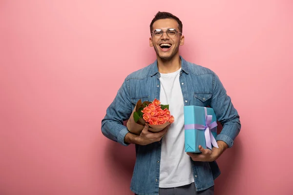 Positiver Mann mit Blumenstrauß und Geschenkschachtel mit Blick in die Kamera auf rosa Hintergrund — Stockfoto