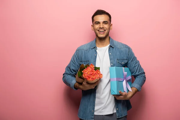 Schöner Mann mit Rosen und Geschenk lächelnd in die Kamera auf rosa Hintergrund — Stockfoto