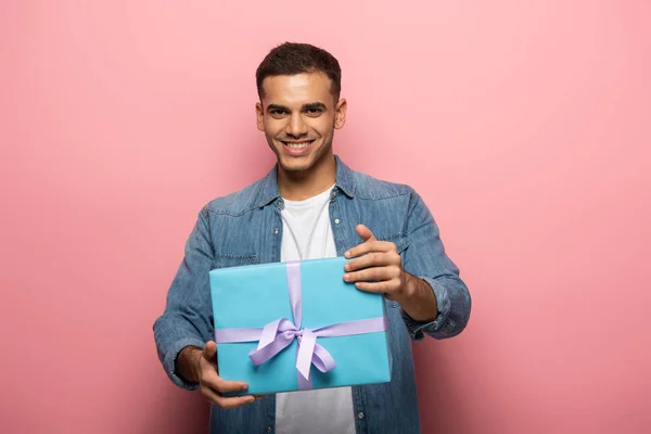 Young man smiling at camera while holding gift on pink background — Stock Photo