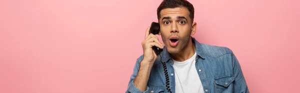 Panorâmica tiro de homem chocado com telefone auscultador olhando para a câmera isolada em rosa — Fotografia de Stock