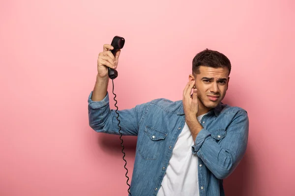 Hombre confuso con auricular telefónico que cierra la oreja sobre fondo rosa - foto de stock