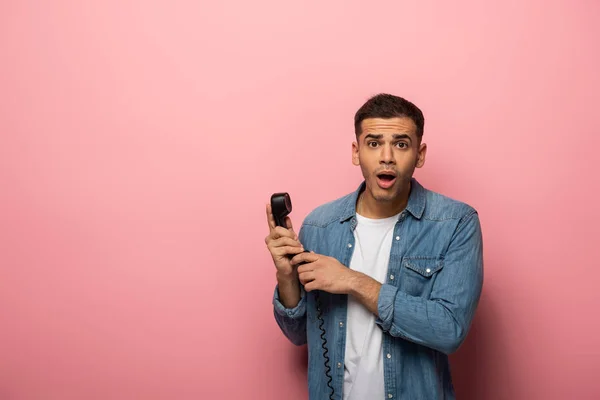 Шокированный молодой человек смотрит в камеру и держит телефон на розовом фоне — стоковое фото