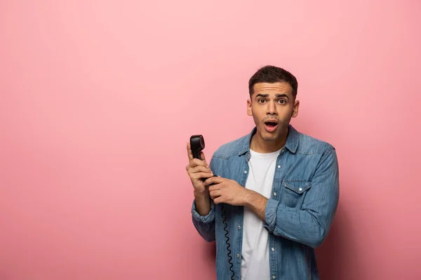 Joven sorprendido mirando a la cámara y sosteniendo el auricular del teléfono sobre fondo rosa - foto de stock