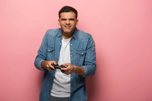 KYIV, UKRAINE - NOVEMBER 12, 2019: Young man with gamepad grimacing at camera on pink background — Stock Photo