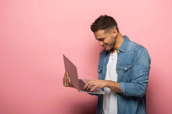 Vista lateral del hombre sonriente utilizando el ordenador portátil sobre fondo rosa - foto de stock
