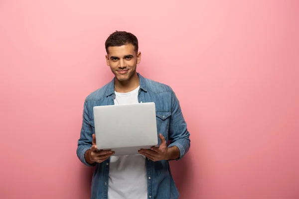 Красивый мужчина держит ноутбук и улыбается в камеру на розовом фоне — стоковое фото