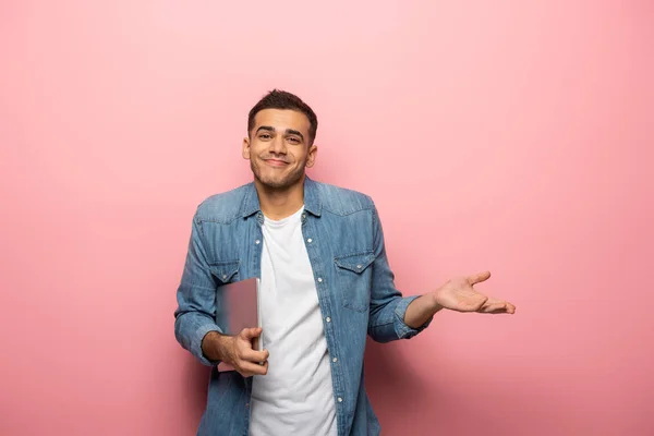 Young smiling man with laptop showing shrug gesture on pink background — Stock Photo