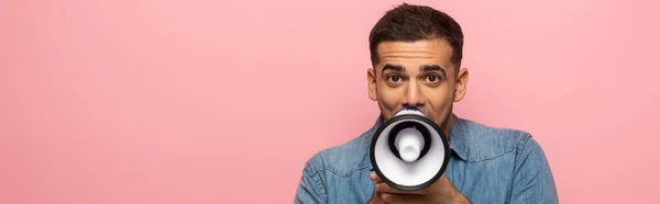 Panoramic shot of man looking at camera while screaming in loudspeaker isolated on pink — Stock Photo