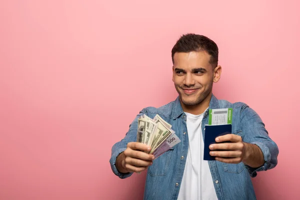 Uomo sorridente che mostra denaro e passaporto con carta d'imbarco su sfondo rosa — Foto stock