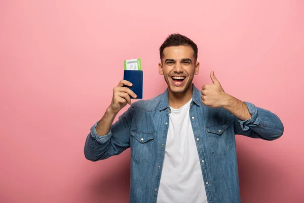 Hombre sonriente sosteniendo pasaporte con tarjeta de embarque y mostrando como signo sobre fondo rosa - foto de stock