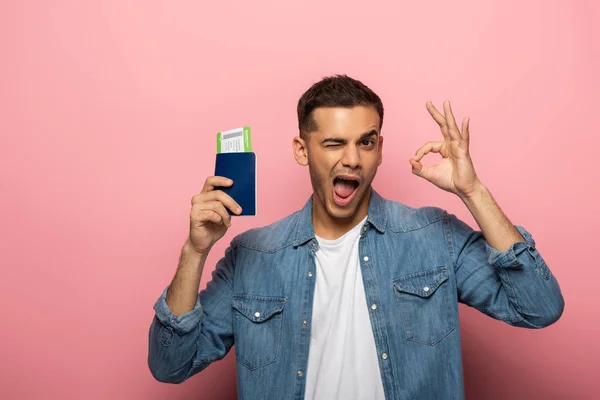 Homme clignant des yeux et montrant un geste correct tout en tenant passeport avec carte d'embarquement sur fond rose — Stock Photo