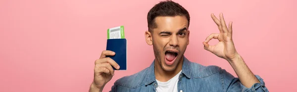 Foto panorámica del hombre con pasaporte y tarjeta de embarque guiñando un ojo y mostrando el signo de bien aislado en rosa - foto de stock