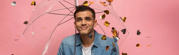 Panorâmica tiro de homem sorridente com guarda-chuva transparente sob queda confete isolado em rosa — Fotografia de Stock
