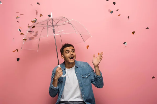Uomo sorpreso con ombrello che punta con mano sotto i coriandoli che cadono su sfondo rosa — Foto stock