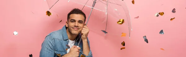 Giovanotto con ombrello sorridente alla macchina fotografica sotto coriandoli a forma di cuore cadenti su sfondo rosa, scatto panoramico — Foto stock