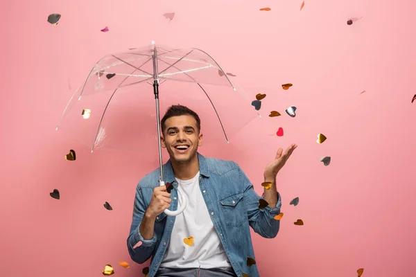 Mann mit Regenschirm lächelt in die Kamera und zeigt mit der Hand unter fallendem Konfetti auf rosa Hintergrund — Stockfoto
