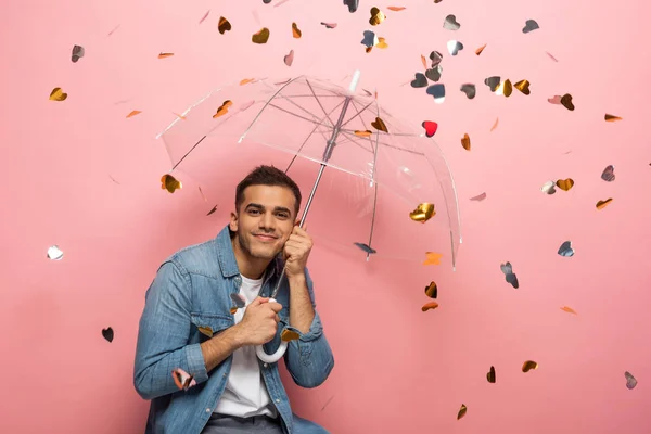 Jeune homme avec parapluie souriant à la caméra sous les confettis en forme de coeur tombant sur fond rose — Photo de stock