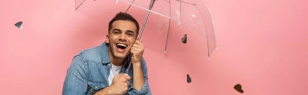 Panoramic shot of cheerful man with transparent umbrella under falling confetti on pink background — Stock Photo