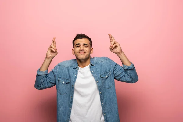 Young man with crossed fingers smiling at camera on pink background — Stock Photo