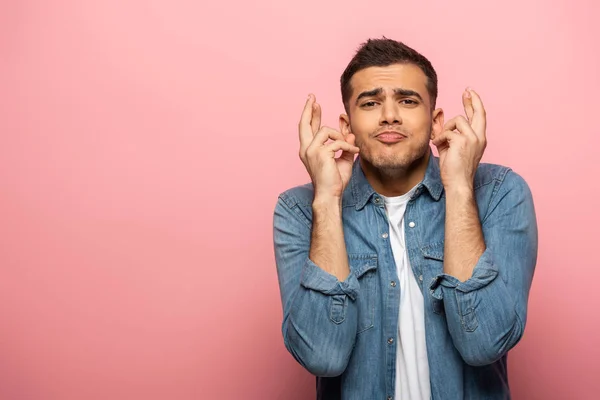 Bonito homem com expressão facial e dedos cruzados olhando para a câmera no fundo rosa — Fotografia de Stock