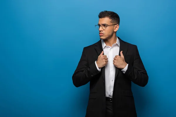 Jovem empresário ajustando jaqueta e olhando para o fundo azul — Fotografia de Stock