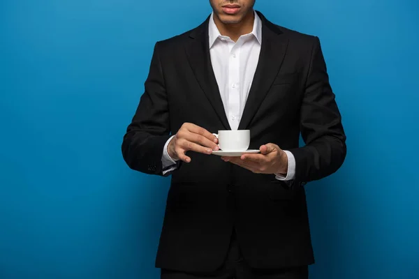 Vista recortada del hombre de negocios sosteniendo taza de café y platillo sobre fondo azul - foto de stock